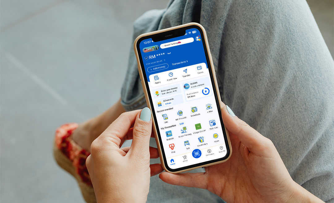 A woman holding a smartphone displaying the homepage of the Touch ’n Go eWallet app, which now supports MDAC submissions.