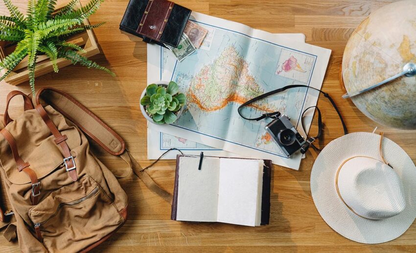 Flat lay of travel essentials including a laptop, paper maps, and a passport on a wooden desk, representing the research phase of planning a group trip.
