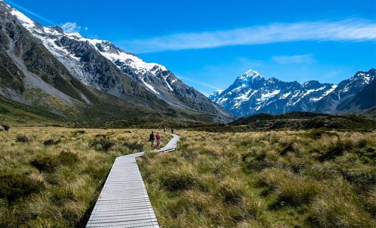 A wooden boardwalk trail winding through alpine grasslands toward a majestic mountain peaks backdrop on the Hooker Valley Track in Mt Cook National Park, New Zealand.