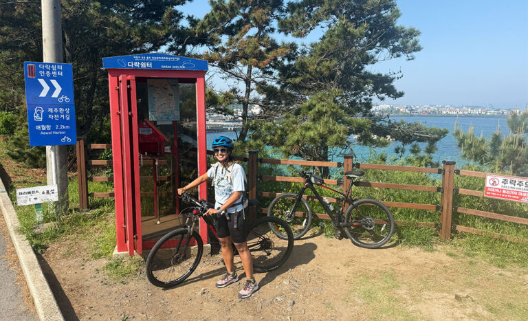 Sofia Shamsunahar posing next to a bright red telephone box stamp station on the Jeju Fantasy Bicycle Path in South Korea.