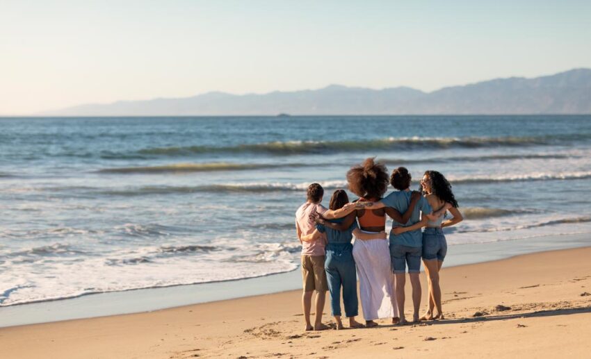 A back-view of five diverse female friends standing on a sandy beach, embracing as they look out together at the ocean waves.
