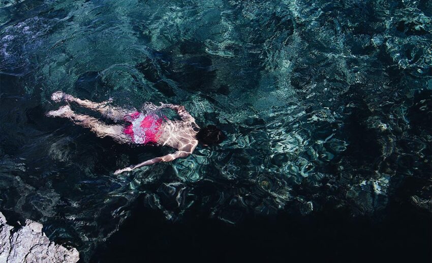 High-angle wide shot of a female swimmer wearing a pink and white swimsuit, navigating clear teal open water.