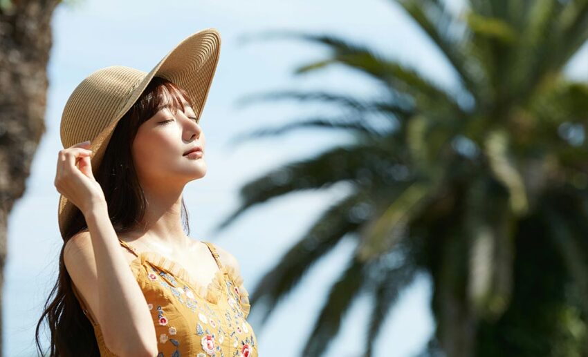 A woman wearing a straw sun hat and sundress looking up peacefully at the warm summer sky with eyes closed.