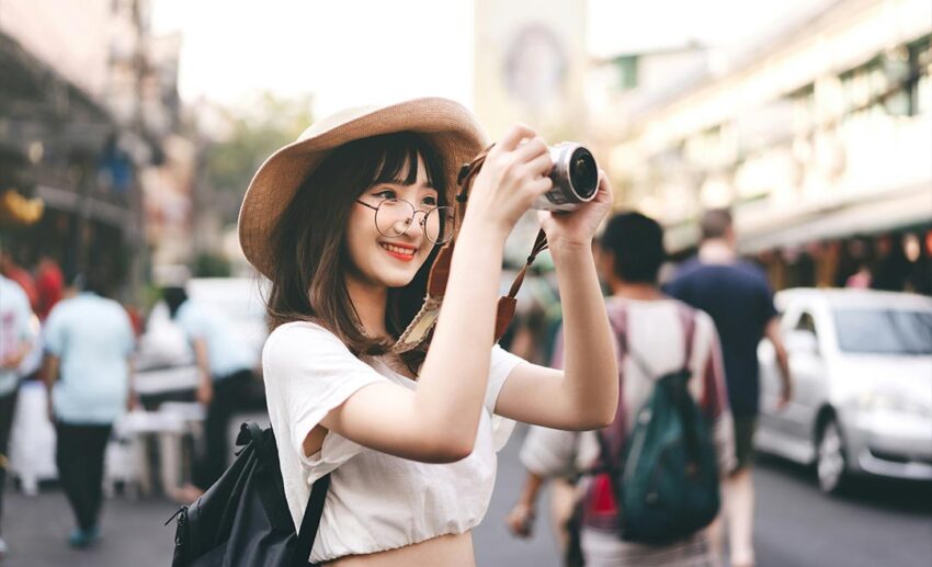 A young Asian solo female backpacker wearing glasses holding a camera and sightseeing on a city street.