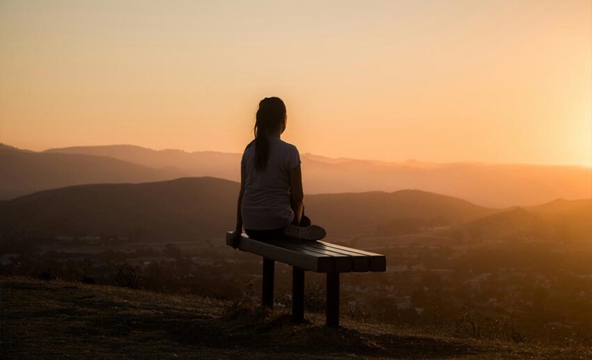 A solo female traveller sitting on a wooden bench overlooking a majestic mountain valley landscape, practising mindful lifestyle design.