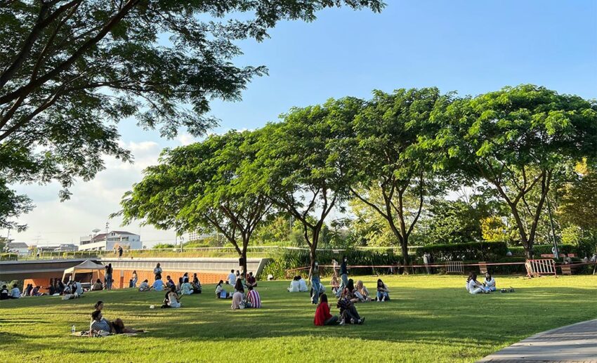 People scattered and relaxing on the lush grass of a vibrant public park in Bangkok, Thailand, on a clear, sunny day.