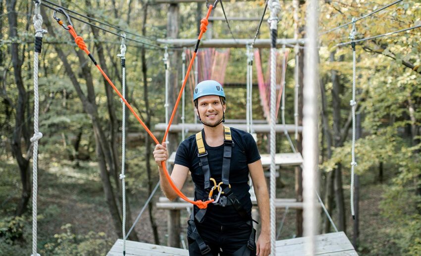 A smiling, professional male climbing instructor wearing a protective safety helmet, a full body harness, and secure carabiners at an outdoor high ropes course.