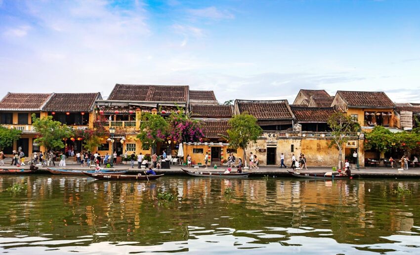 A vibrant daytime view of the historic Hoi An waterfront featuring traditional wooden boats moored next to classic yellow French-colonial architecture in Quang Nam Province, Vietnam.