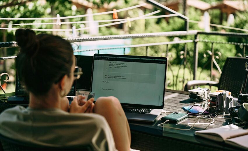 Rear view of a female remote worker sitting at an outdoor terrace table with computing gear, working independently while travelling in Çıralı, Antalya, Turkey.
