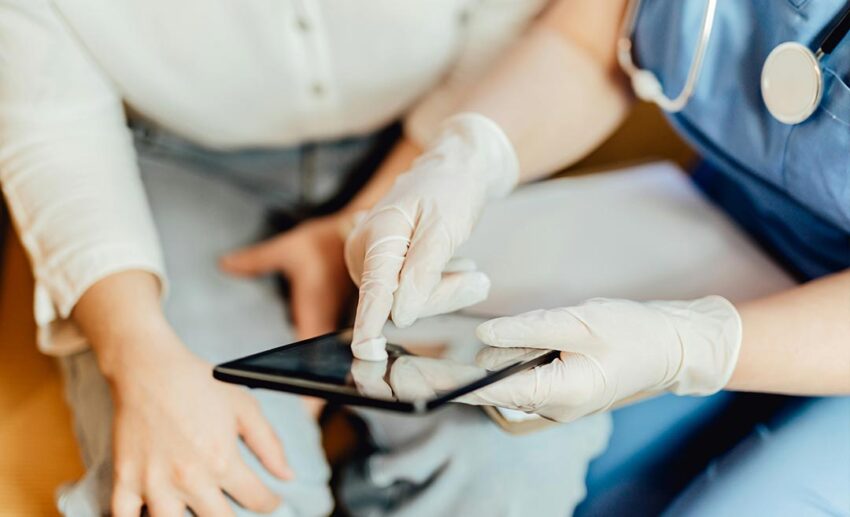 A medical healthcare professional in blue hospital scrubs and white protective gloves holding a smartphone, representing European healthcare systems.