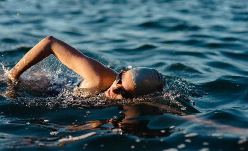 Close-up side view of a focused female open water swimmer wearing a protective swim cap and goggles in the water.