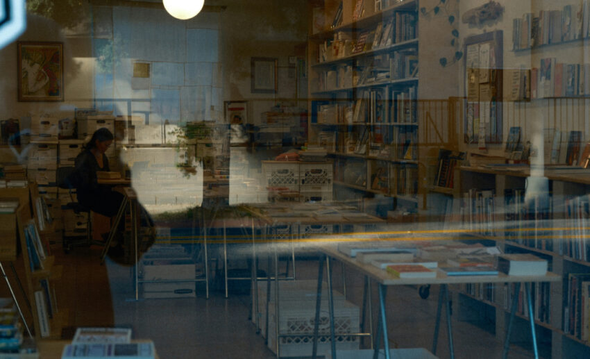 A woman sitting quietly and reading a book inside a cosy, empty bookstore, viewed through the exterior glass window.