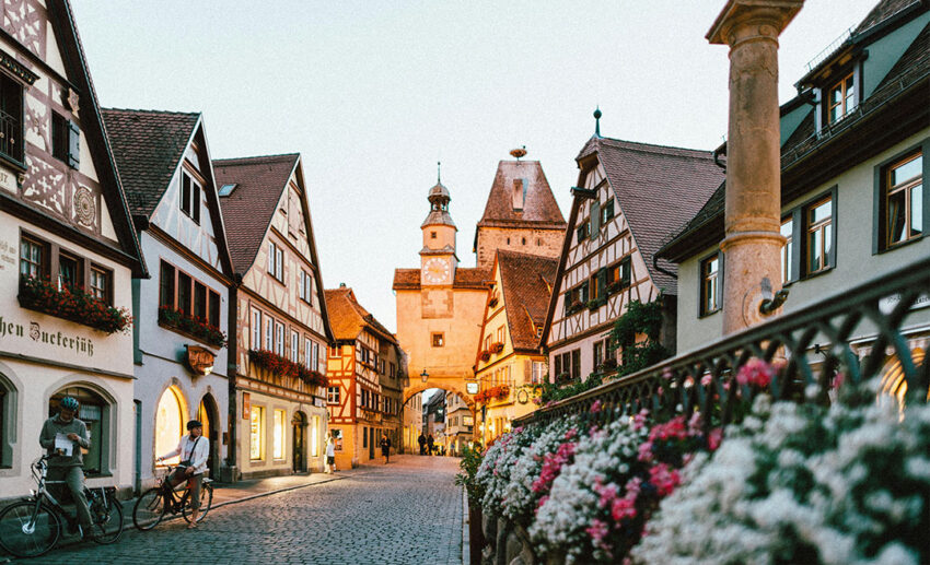Beautiful pink and white flowers blooming on a metal fence with fairy-tale German architecture and a historic stone tower in Rothenburg ob der Tauber in the background.