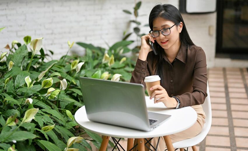 A remote female worker using her phone and laptop outdoors, representing a digital nomad lifestyle while living abroad as a Malaysian.
