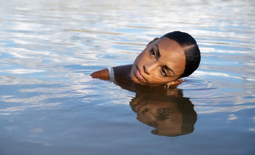 A calm, eye-level shot of a woman submerged in smooth open water up to her neck, looking out toward the horizon at sunset.