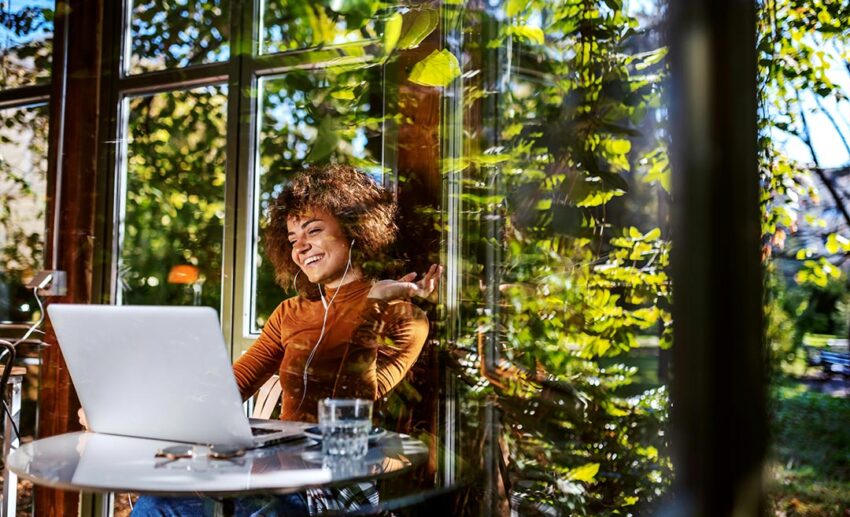 A professional remote worker with curly hair smiling during a video call on her laptop inside a bright, modern European cafe.