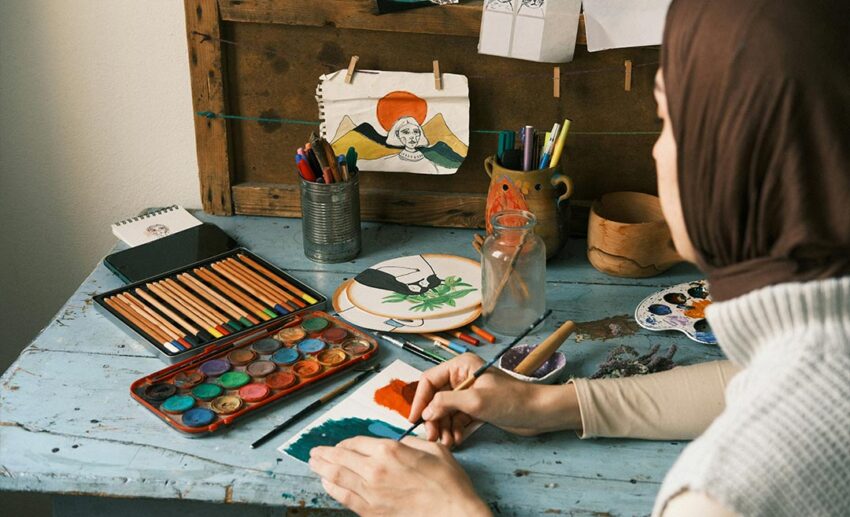 A relaxed woman exploring a creative hobby by watercolour painting at a table filled with art supplies during a mid-career sabbatical.