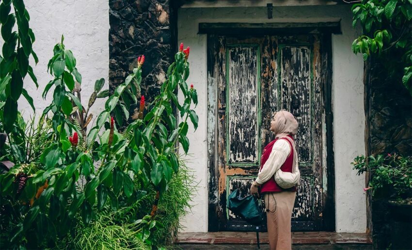 A profile view of a woman standing thoughtfully by an open arched doorway framed by lush green tropical plants.