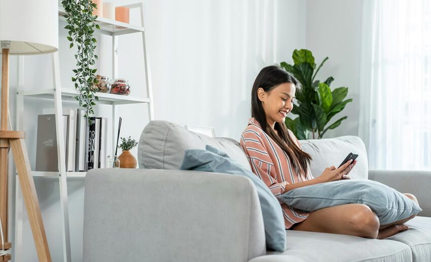A woman relaxing on a living room sofa while using her mobile phone to check local amenities inside a rented apartment abroad.