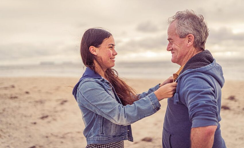 Adult daughter and elderly father laughing and hugging on a sunny beach during family multigenerational travel