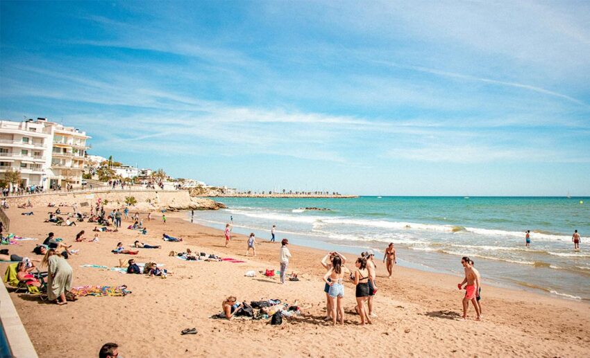Groups of people unwinding and walking along a wide sandy beach under a bright sky during the daytime.