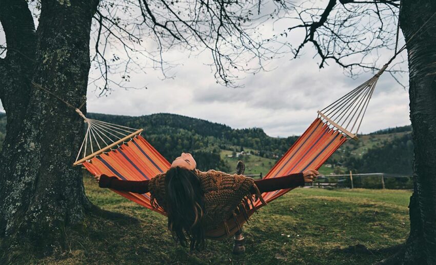 A calm view of a woman resting in a hammock tied under a tree overlooking a peaceful mountain valley landscape.