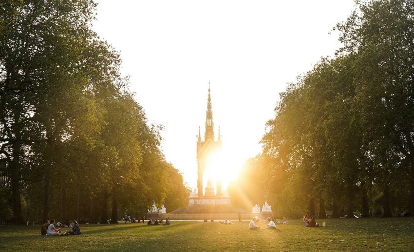 People walking and sitting in groups near a fountain in front of the ornate Gothic Revival Prince Albert Memorial in Hyde Park London during an autumn sunset.