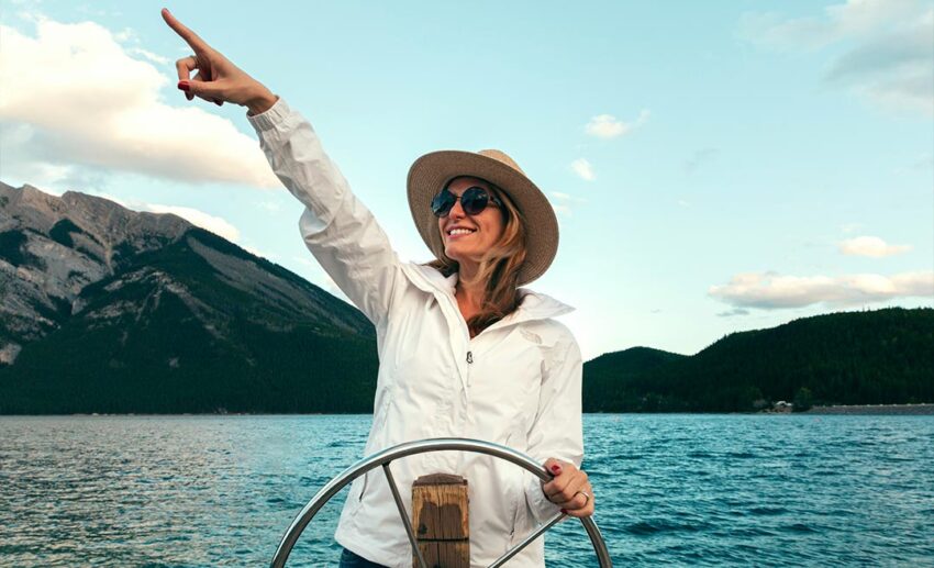 A smiling independent woman confidently steering a boat by the wheel while looking out toward the horizon on a sunny day.