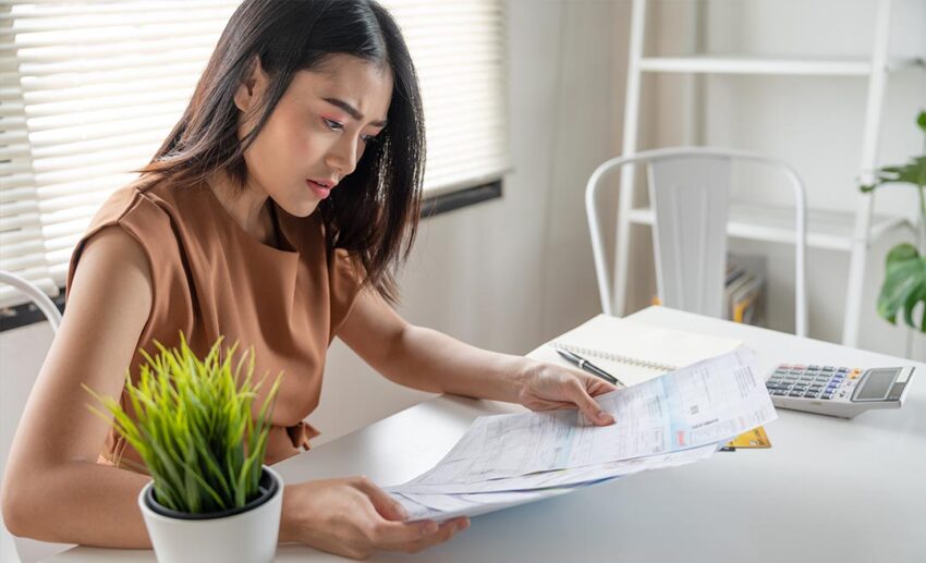 A stressed woman sitting at a desk, looking down anxiously at a spread of paper financial bills and monthly expense invoices.