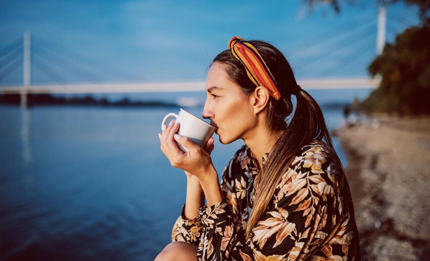 Side profile of a woman wearing a floral summer dress and headband, sitting on a coastal shore while quietly drinking from a mug.
