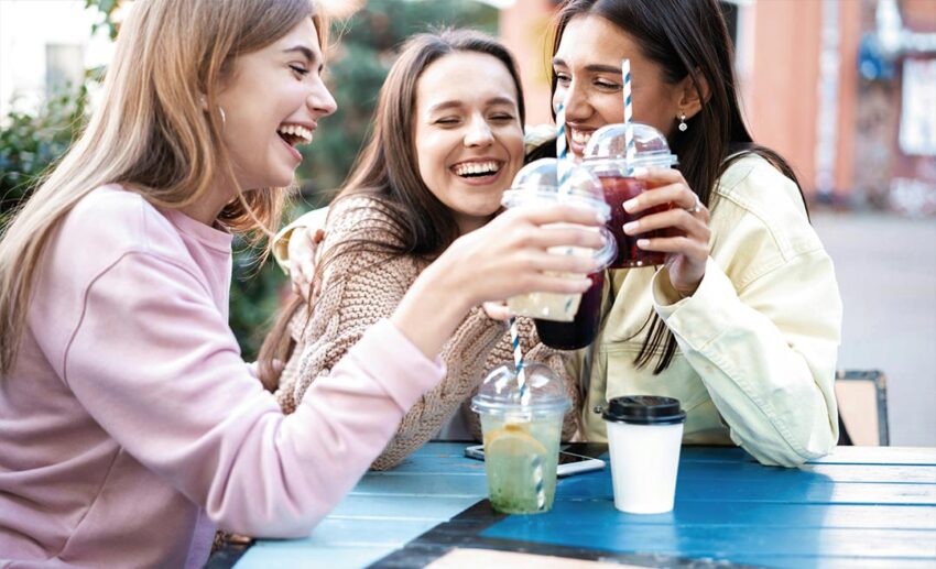 Three cheerful female friends laughing together and drinking colourful cocktails at an outdoor holiday cafe.
