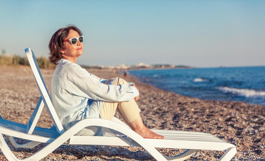 A relaxed middle-aged woman sitting comfortably on a beach lounge chair, watching a golden ocean sunset during a slow travel trip.