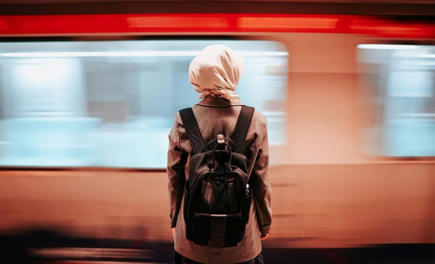 A female traveller with a backpack standing still on a station platform as a blurred train speeds past her.