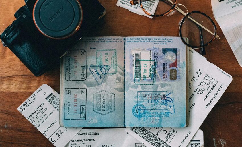 An open passport displaying international immigration stamps, laid flat alongside eyeglasses, a camera, and airline boarding slips for an extended holiday.