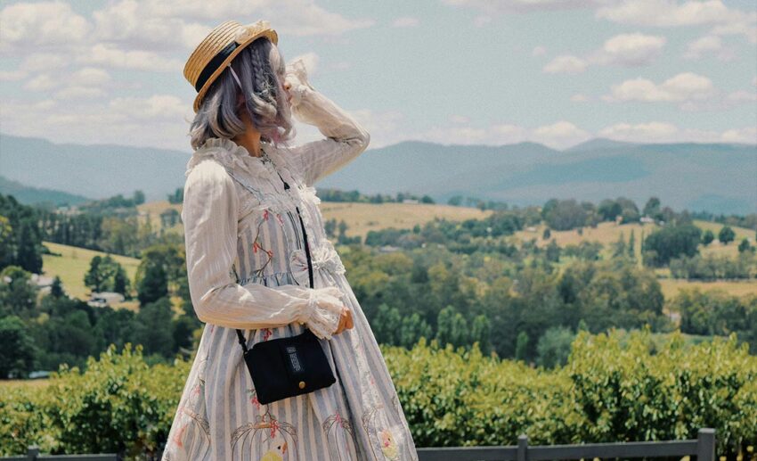 A woman wearing a sun hat and a long dress standing alone on top of a grassy hill, looking out at a vast landscape.
