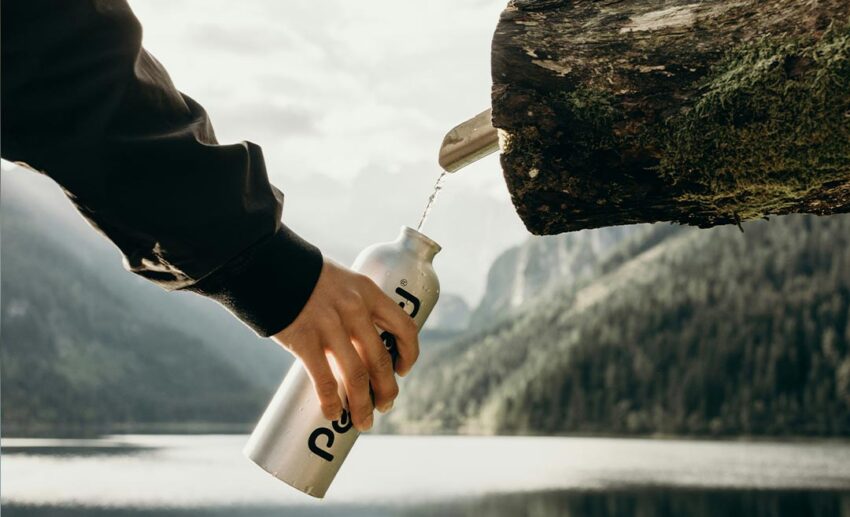 Close-up of a traveller holding a clear reusable water bottle directly underneath a flowing stream of clean, fresh water.