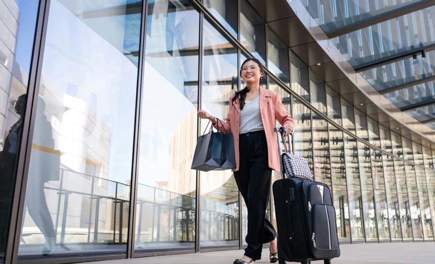 A young Malaysian female traveller pulling a wheeled suitcase and walking through a bright modern airport terminal.