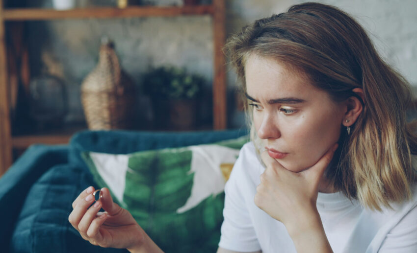 A thoughtful young woman sitting alone at home, holding a ring and looking down in contemplation during a difficult relationship crossroads.