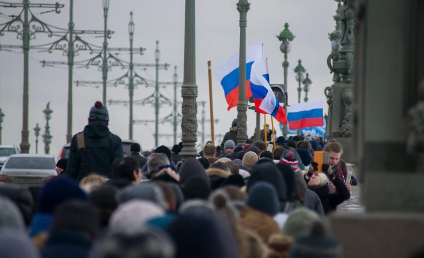 A massive public protest march filled with crowds holding a large black commemorative banner crossing the Trinity Bridge in Saint Petersburg, Russia.