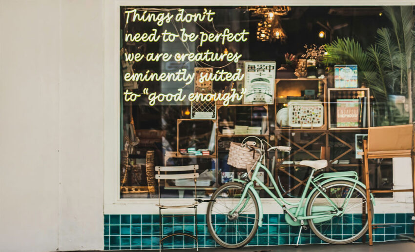 The exterior glass window of a trendy cafe in Tiong Bahru, Singapore, displaying a philosophical quote about being suited to "good enough," with an old-timey green bicycle leaning against the glass.