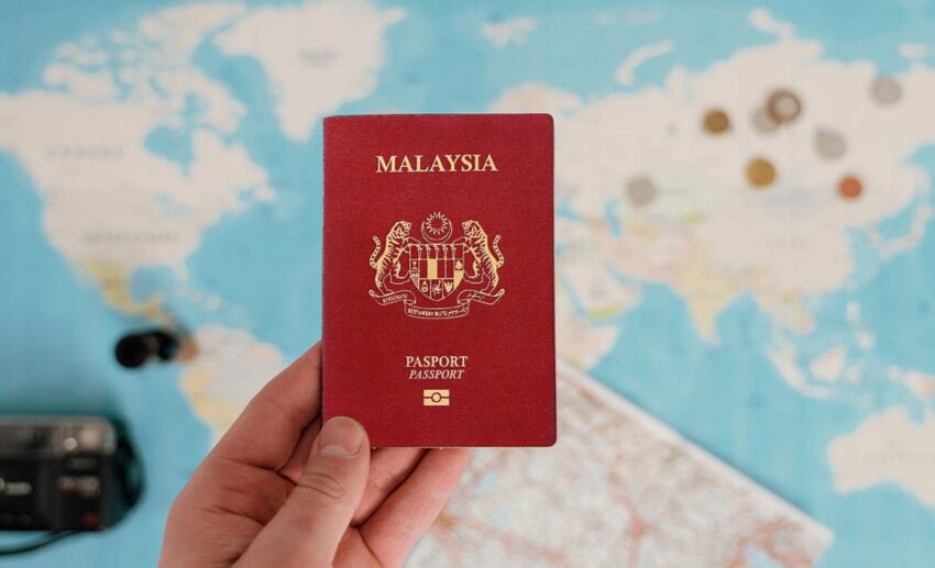 A close-up of a person's hand holding a red Malaysian passport over an open world map during travel planning.