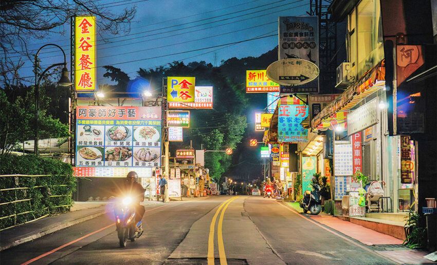 A tranquil night scene in the Guguan Hot Springs resort area of Taichung, Taiwan, with luminous traditional Chinese signs glowing against a twilight mountain backdrop as a lone scooter drives past.