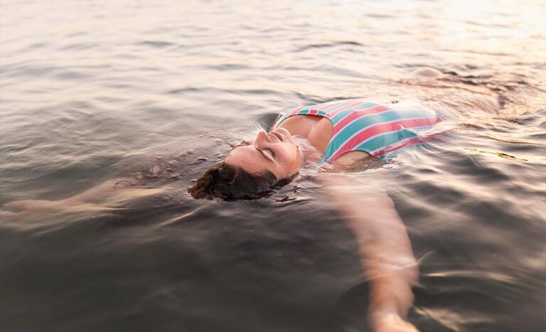 A serene, wide shot of a woman floating face up in the calm sea, completely relaxed and buoyant in the open water.