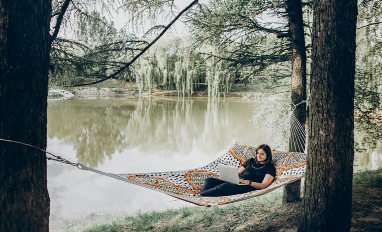 A woman working remotely on a laptop while relaxing in a hammock at a sunny park during a micro retirement career break.