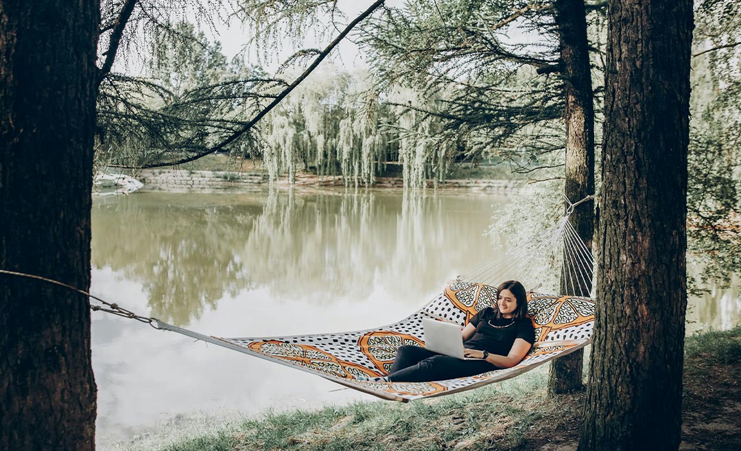 A woman working remotely on a laptop while relaxing in a hammock at a sunny park during a micro retirement career break.