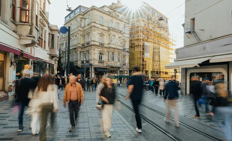 A long exposure photo of a crowded city street with blurred motion of busy people moving fast past modern urban buildings.