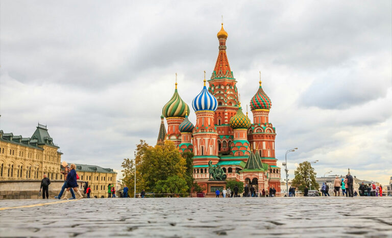A crowd of tourists walking along the cobblestone streets of Moscow, with the colourful domes of Saint Basil's Cathedral visible in the background.