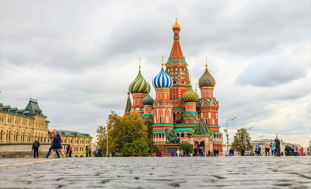 A crowd of tourists walking along the cobblestone streets of Moscow, with the colourful domes of Saint Basil's Cathedral visible in the background.