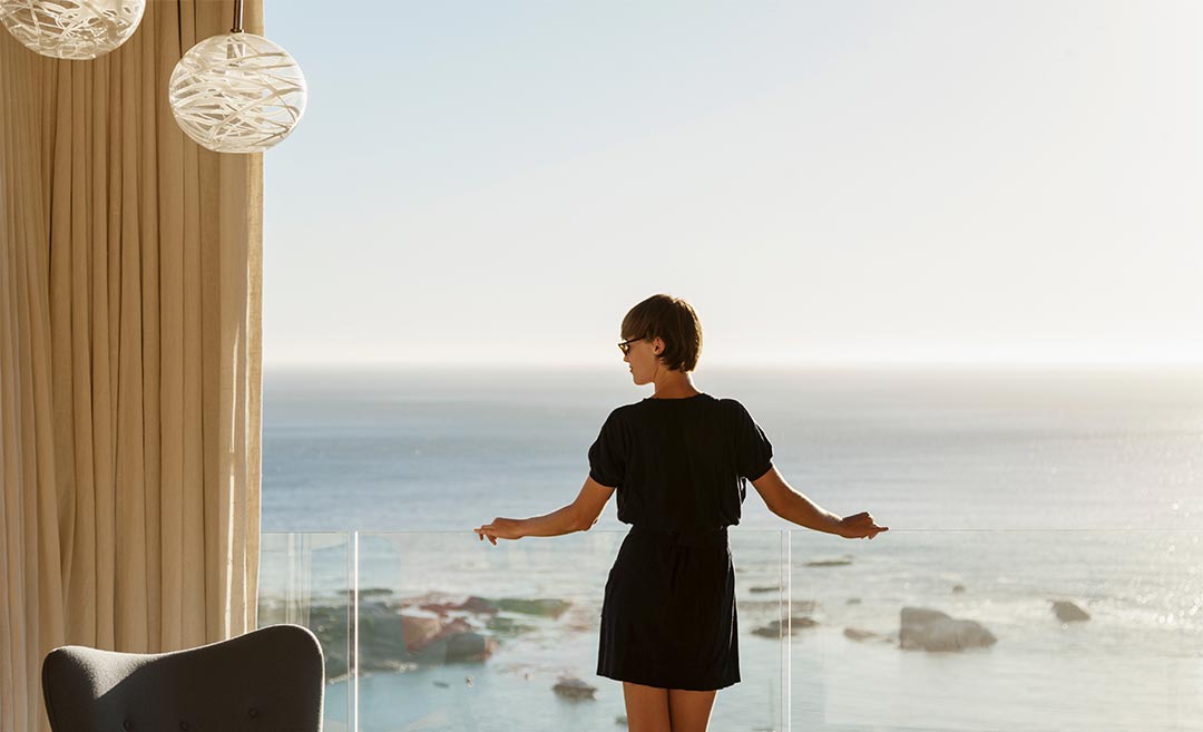 A female traveller standing at an open balcony railing, looking out over a wide, blue tropical ocean during a long-term trip.
