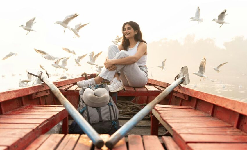 A solo female traveller sitting peacefully in a wooden boat on the water, surrounded by flying birds at dawn.